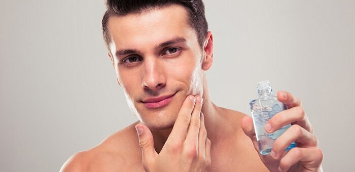 a young attractive man applying aftershave on his face and holding an aftershave bottle in his other hand