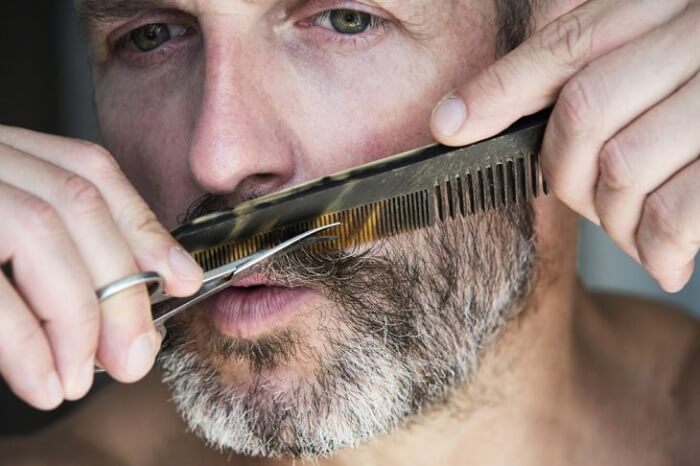 man with grey hair trims his mustache and beard with scissors and comb